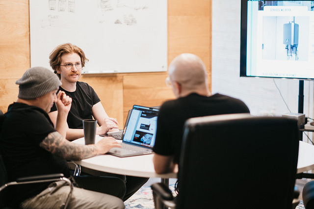 Three team members collaborate around a table in a modern office, reviewing a technical diagram displayed on-screen. Laptops are open for active discussion, and a whiteboard provides additional context for their in-house strategy session.