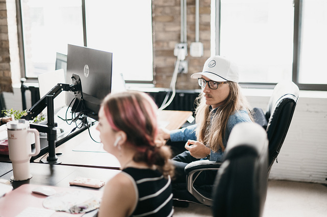 A man with long hair and a white cap sits at a desk, reviewing in-house vs. outsourced marketing strategies with a woman sporting pink-highlighted hair and earbuds. They collaborate in a modern, well-lit office—illustrating Hook Agency's commitment to creative, results-driven teamwork.