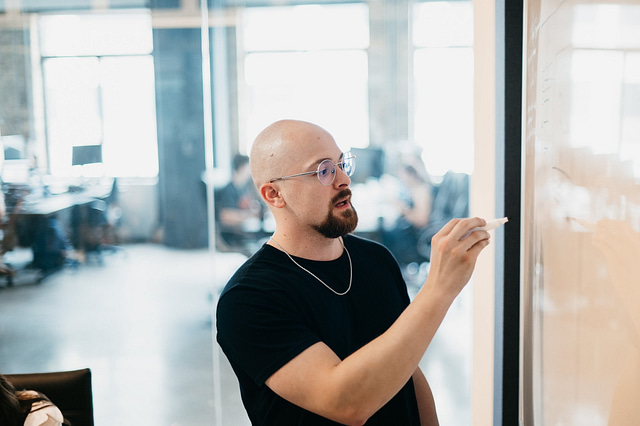 A man with a shaved head and glasses outlines the key differences between rebranding and digital marketing on a whiteboard in a modern office, while colleagues work in the background.