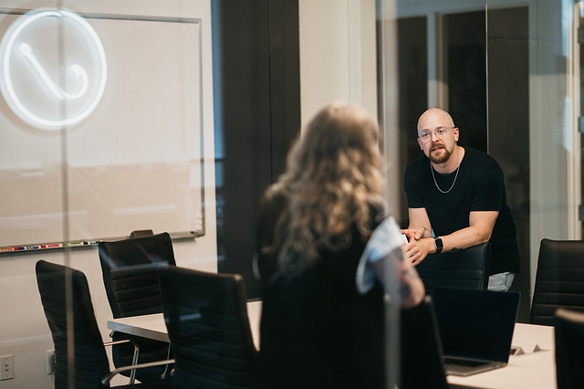 A man and woman engage in a focused discussion on digital advertising strategies in a modern conference room. The setting features black chairs, a glass wall, a laptop on the table, and a whiteboard in the background—reflecting Hook Agency’s commitment to collaborative, results-driven marketing solutions.