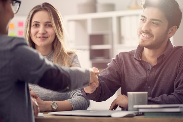 A man and woman sit at a table, smiling, exemplifying customer appreciation. The man extends a handshake to another individual, whose back faces the camera. A laptop and coffee mug on the table suggest a discussion on successful referrals in an inviting office setting.