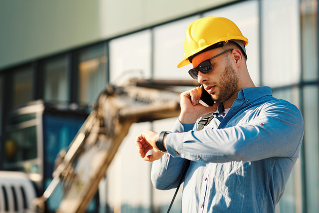 A contractor, identifiable by his yellow hard hat and sunglasses, stands confidently in front of a building and construction equipment. He checks his watch while speaking on the phone to ensure the day's work starts promptly as scheduled.