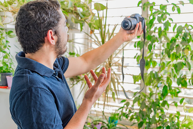 On Hook Agency's website, a bearded man with dark hair stands indoors, clad in a dark blue shirt. He shares valuable roofing tips through the window adorned with plants. Captured in bright streaming light, he offers insightful demonstrations to his social media followers.