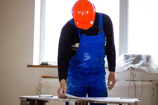 A general contractor, clad in a bright orange hard hat and blue overalls, meticulously inspects a white material indoors. Surrounded by tools of renovation—closed windows and covered objects—the scene reflects the precision and expertise inherent in the contractor's craft.