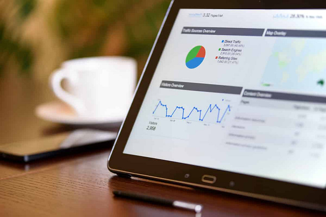 A tablet on a table displays a digital dashboard with charts and graphs, including a traffic source pie chart and a line graph of visitor data, illustrating the impact of SEO pricing. In the background, a smartphone, pen, and coffee cup complete the scene.