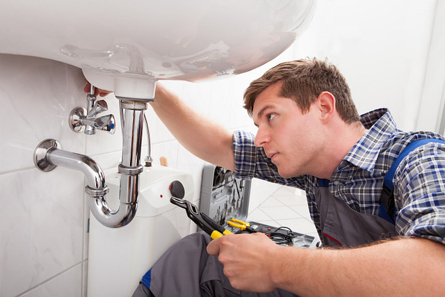 A checkered-shirted plumber expertly tightens a pipe under the sink, exemplifying "Your Trusted Partner in Plumbing." The bright bathroom, featuring white tiles and a glimpse of the washing machine, highlights our exceptional plumbing services.