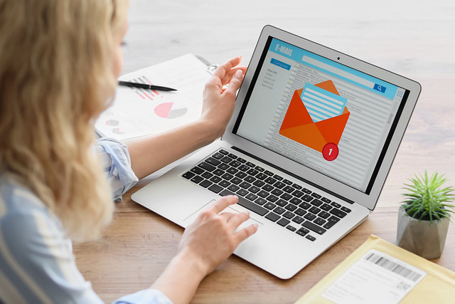 On a wooden desk, a person reviews a laptop displaying an email notification. Accompanying the setup are a small plant, yellow envelope, and various documents. The email reveals an open red envelope icon with a blue letter inside, announcing exclusive commercial roofing deals via cold email.