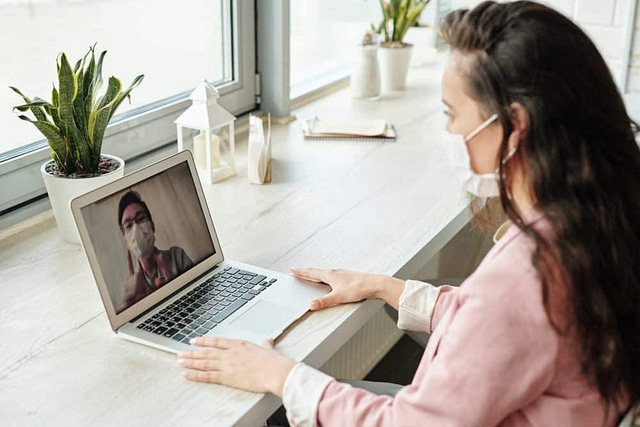 On Hook Agency's website: During the COVID-19 pandemic, a woman wearing a face mask participates in a video call with masked contractors. Her laptop signifies the current climate of caution. The workspace is illuminated by natural light, featuring potted plants and a closed notebook.