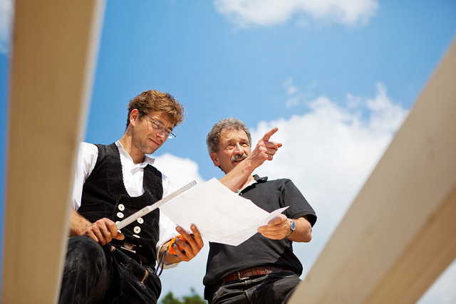 At a construction site, two men review plans to ensure accountability. One man, in a safety vest, holds a measuring tool while the other directs attention toward the future build. They stand surrounded by wooden beams under a clear blue sky.