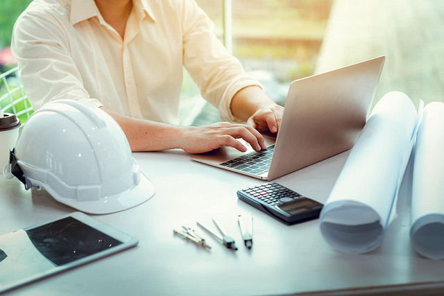 On Hook Agency's website, a professional in a white shirt is actively engaged on a laptop at a desk. The workspace is equipped with essential tools including a tablet, hard hat, rolled-up blueprints, calculator, and drafting instruments. This well-lit setting emphasizes precision in design and engineering tasks, likely integrating advanced elements of website design to boost functionality.