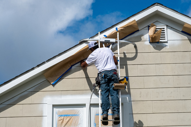 A man on a ladder expertly paints the exterior of a house, demonstrating precise exterior remodeling techniques. Masking paper is strategically placed to shield specific areas, ensuring a flawless finish. The partly cloudy sky indicates ideal conditions for this specialized work.
