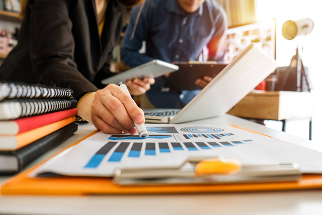 A person highlights a bar graph on a clipboard with a pen, demonstrating effective strategies for growth in the construction industry. Another team member engages with a tablet, surrounded by notebooks and a laptop on the desk. The bright lighting underscores the collaborative atmosphere at Hook Agency.