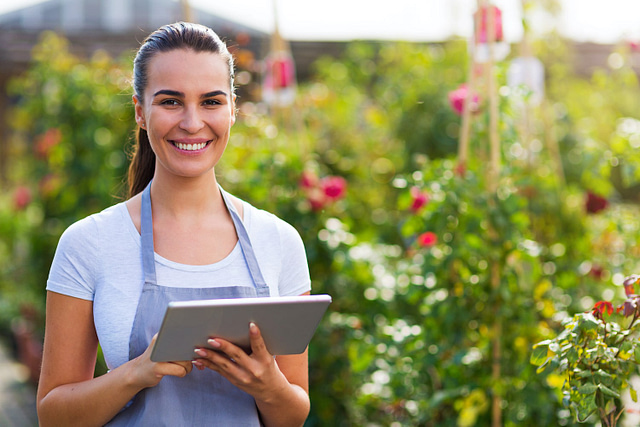 A woman in an apron stands confidently in a lush garden, holding a tablet amid blooming flowers and greenery. This picturesque scene exemplifies vibrant and effective landscape marketing, infused with warmth and energy.