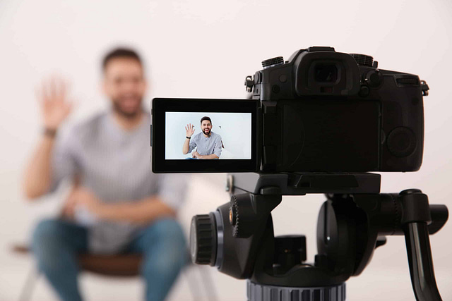 A man sits confidently on a chair, smiling and waving at the camera, poised to present his latest YouTube video. The camera's flip screen prominently displays his image against a simple, light background.