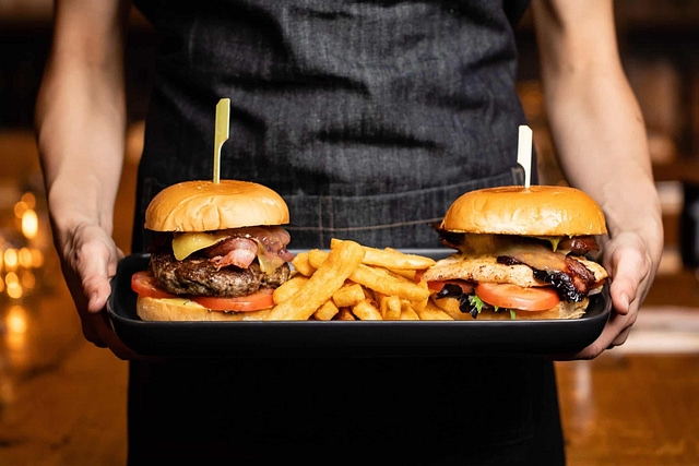 A person confidently presents a tray featuring two of Minneapolis's finest burgers, complete with thick-cut fries. Each burger is expertly assembled with fresh tomatoes, crisp lettuce, and melted cheese, secured by skewers. The server wears a dark apron emblematic of the city's vibrant restaurant scene.