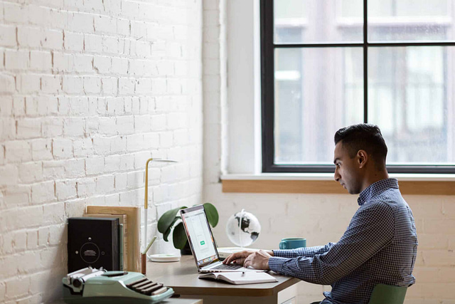On Hook Agency's website, a focused individual works diligently at a desk positioned by a window. This clutter-free workspace is optimized for productivity, featuring essential items like a lamp, a book, a green typewriter, and a globe. The well-lit room boasts white brick walls and is enhanced by natural light streaming through the large window.