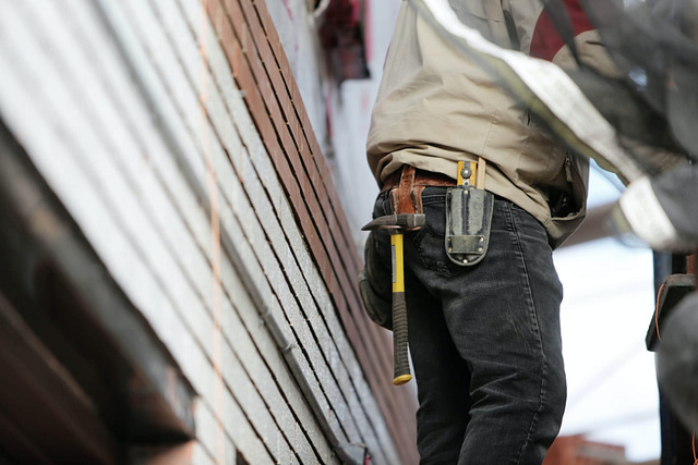 A roofing specialist, equipped with a tool belt containing a hammer and essential instruments, stands beside a stack of bricks. With focused attention, they skillfully update the roofing elements of the building, partially concealed by a tarp.