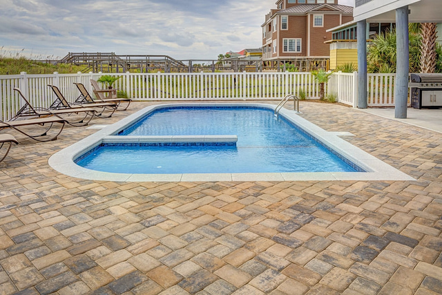Experience the excellence of Pool Installer Marketing with a pristine rectangular swimming pool, paired with a stylish paved patio. Relax on the lounge chairs to the left or fire up the gas grill to the right. In the backdrop, a charming wooden house stands under a cloudy sky, all enclosed by a classic white fence.