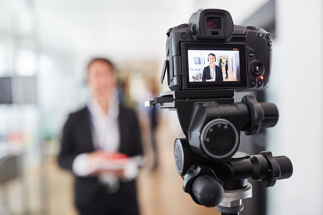 A digital camera mounted on a tripod captures a professional marketing presentation indoors. The screen shows an individual in formal attire, holding a red folder, delivering step-by-step insights. A softly blurred background enhances the professional ambiance.