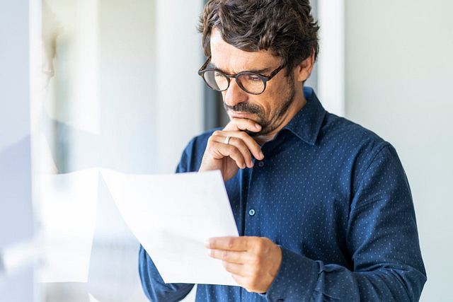 On Hook Agency's website, a focused man in glasses and a dark shirt embodies the CEO mindset as he studies a document. His hand on his chin, exuding deep contemplation. The softly blurred background underscores the intensity of this decisive moment.