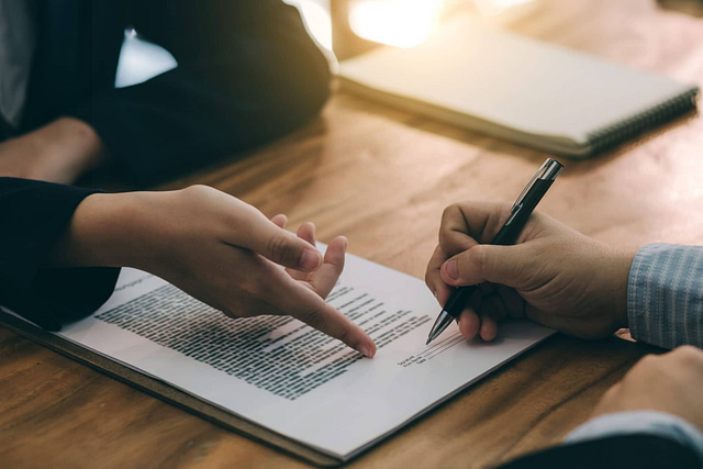 Two professionals, dressed in business attire and seated at a wooden table, finalize a "Roofing Estimates" document. Sunlight filters through, illuminating the scene and emphasizing a professional atmosphere.