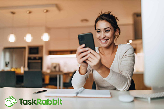 Task Rabbit for Contractors Handyman Home Services Reviews: A smiling woman uses her smartphone at a desk in a modern kitchen, likely checking TaskRabbit reviews. A notebook and pen lie in front of her. The kitchen features pendant lighting and a warm color scheme, with the TaskRabbit logo in the bottom left corner.