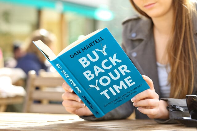 A reader is seated at an outdoor wooden table, engrossed in "Buy Back Your Time" by Dan Martell. The striking blue cover draws attention as they explore strategies to regain control and escape burnout, set against a softly blurred backdrop that enhances a sense of tranquility.