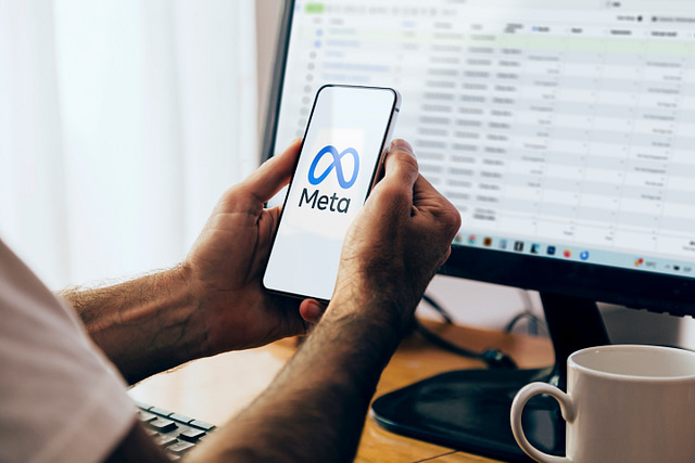 On Hook Agency's website, an individual confronts unauthorized Facebook charges while holding a smartphone with the Meta logo. In the background, a computer monitor displays a detailed spreadsheet tracking these discrepancies. A white coffee mug sits nearby, fueling focus and determination.