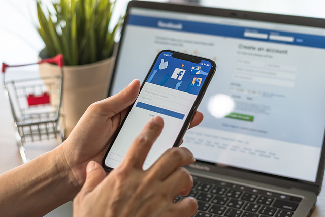 A smartphone displays the Facebook login screen, with a laptop in the background also open to Facebook’s login and sign-up page—representing effective management of home services Facebook ads. A small shopping cart and plant complete the workspace setting.