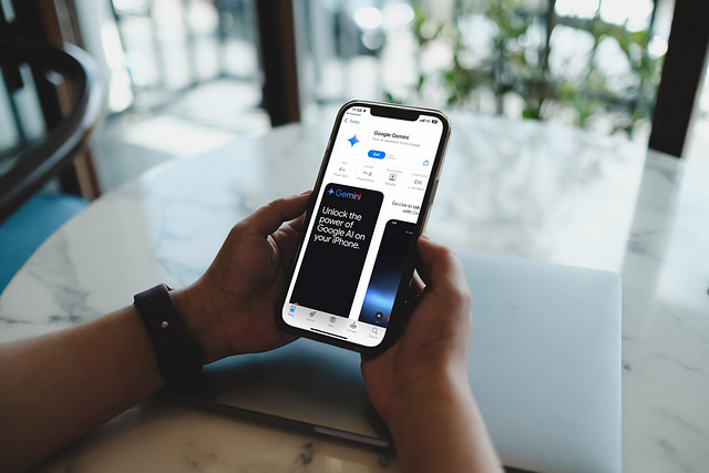 A person holds an iPhone open to the Google Gemini app page on the App Store, showcasing features like Generative Engine Optimization (GEO) designed for home service businesses. In the background, a blurred laptop sits on a marble table near a window with greenery outside.
