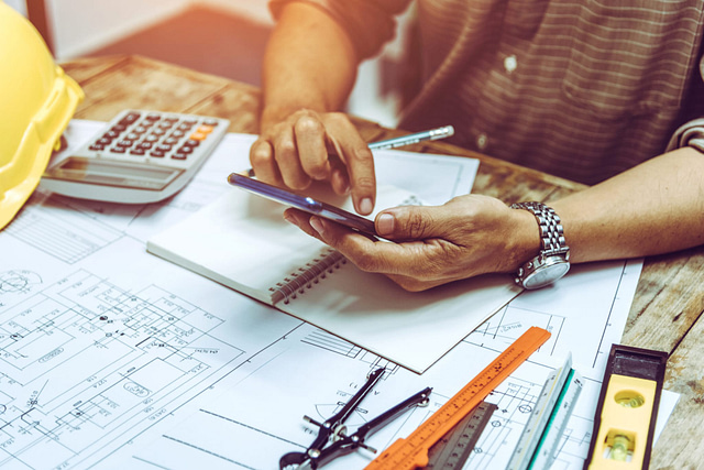 A contractor, equipped with a smartphone, sits at a desk surrounded by engineering blueprints, a calculator, a yellow hard hat, notebook, compass, and drafting tools—actively optimizing their construction or engineering project.