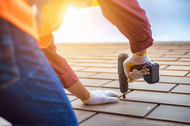 As the sun sets, a professional in gloves and a safety vest diligently repairs a roof with a power drill. This ideal summer moment can be leveraged to boost your online presence. Check our site for effective roofer marketing tips at Hook Agency.