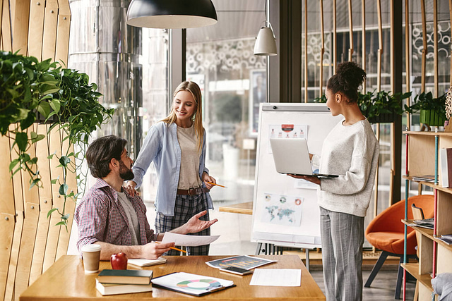 Three professionals collaborate seamlessly in a modern office. One reviews documents at the table, another engages with a laptop, and the third presents data on a flip chart. This well-lit, contemporary space captures the dynamic nature of marketing agencies.