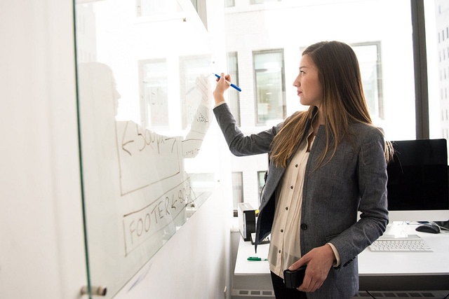 A woman in a gray blazer, likely a Marketing Manager, writes on a whiteboard with a blue marker in an office setting. She appears focused, illustrating a flowchart or diagram. A computer and window are visible in the bright room.