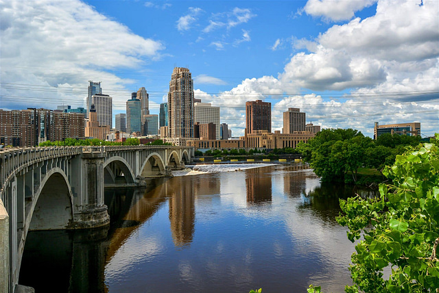 Experience the stunning Minneapolis skyline as it reflects off the tranquil river under a bright blue sky. A prominent bridge spans the water, while lush greenery lines the banks, creating an ideal backdrop for showcasing weekend adventures in this dynamic city.