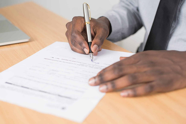 On a wooden desk, a person wearing a gray shirt and black tie confidently signs a construction contract with a silver pen. A laptop, perfect for accessing top construction contracts, is partially visible in the background.