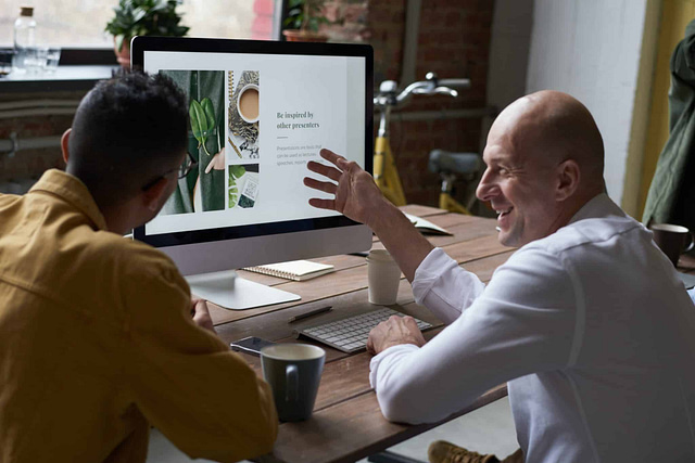 Two individuals sit at a wooden table, intently focused on a computer screen showcasing a plant-themed presentation. One person, assuming the leadership role, gestures toward the screen as they engage in a dynamic discussion. Nearby coffee cups underscore their commitment to productive collaboration.