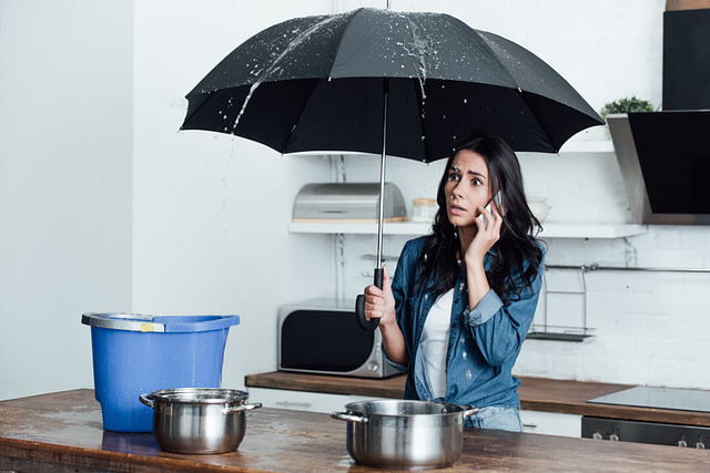 A woman stands in her kitchen with an open black umbrella, speaking on the phone as water drips from the ceiling into containers scattered on the table and floor. Her worried expression suggests she's considering urgent water damage restoration to address the leak.