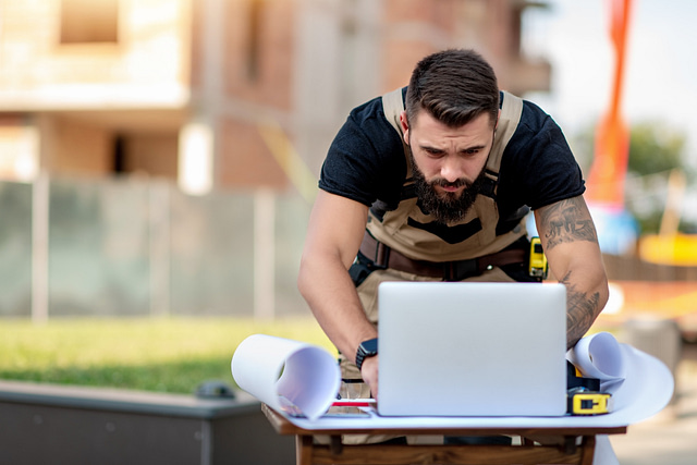 A bearded, tattooed construction worker in a tool belt and work clothes reviews blueprints and works on a laptop at an active construction site.
