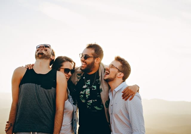 Four friends gather outdoors under a bright sky, exuding happiness and humor. One sports sunglasses and a tank top, another wears a patterned shirt, while two are in casual attire. They are close-knit and jovial, embodying the essence of camaraderie.