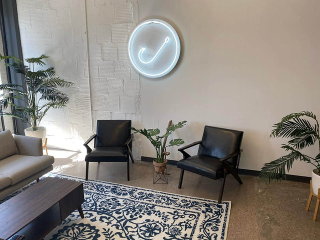 Enhance your leads in 2024 with our inviting modern waiting area. Featuring two sleek black chairs, a gray sofa, and a wooden coffee table on a decorative rug, the space is accented by large potted plants that bring in natural greenery. A minimalist neon sign reading "Better Your Leads" sets a cozy and professional tone.