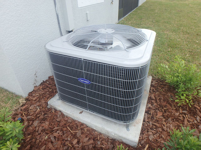A top HVAC brand's outdoor air conditioning unit is securely positioned on a concrete pad, surrounded by wood mulch and lush greenery. This metallic unit, designed with horizontal vents and a top-mounted fan, complements the light-colored walls of the adjacent building.