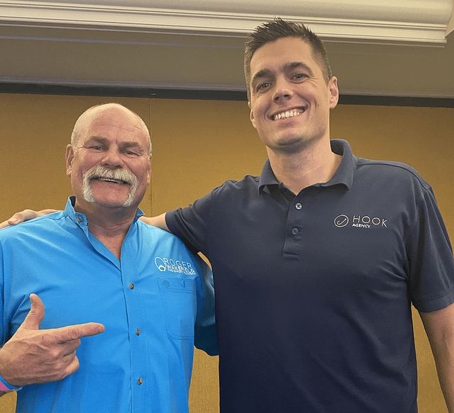 On Hook Agency's website, two charismatic influencers stand side by side against a beige wall. The man on the left confidently showcases his blue shirt adorned with text, while his counterpart proudly wears a dark shirt featuring both a logo and text. Their dynamic presence radiates creativity and charm.