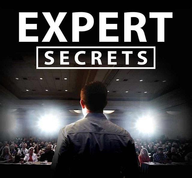 On stage, a speaker faces a large audience, backlit by bright lights. The bold text "EXPERT SECRETS" is prominently displayed above. Attendees seated at tables listen intently, highlighting an environment more akin to a seminar or conference than merely a book review.