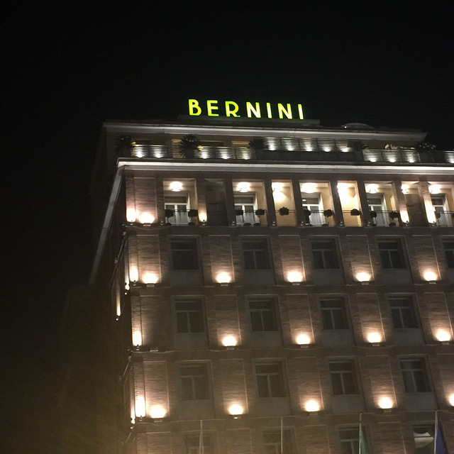 A tall, multi-story building with the word "Bernini" illuminated in green typography atop stands proudly. The facade is artfully lit, highlighting its rows of windows and balconies against the night sky, reminiscent of Italy's architectural elegance.