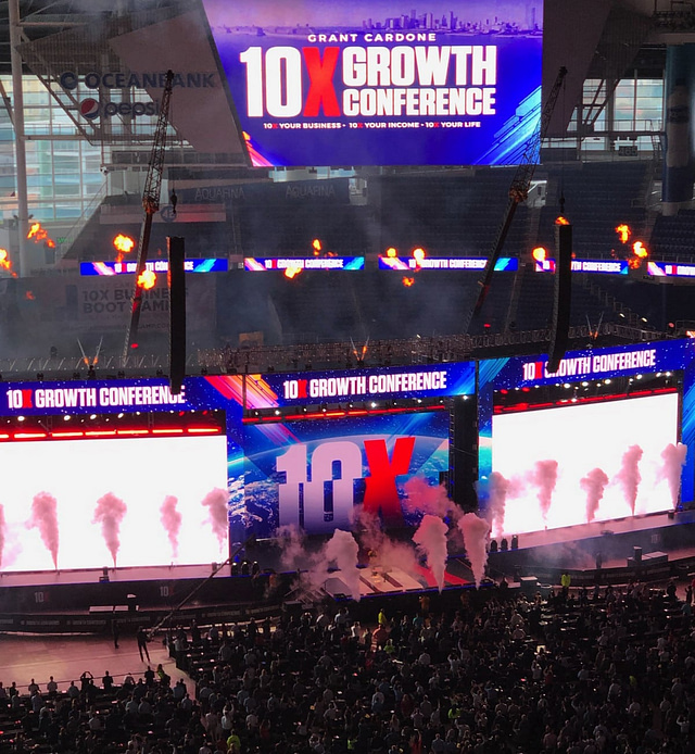 A packed stadium hosts the 10X Growth Conference 2019, featuring bright lights, fireworks, and vivid screens displaying "10X." The stage is surrounded by smoke effects as a lively audience looks on, with Ocean Bank's name visible on the walls. Energetic atmosphere and colorful visuals create an unforgettable recap.