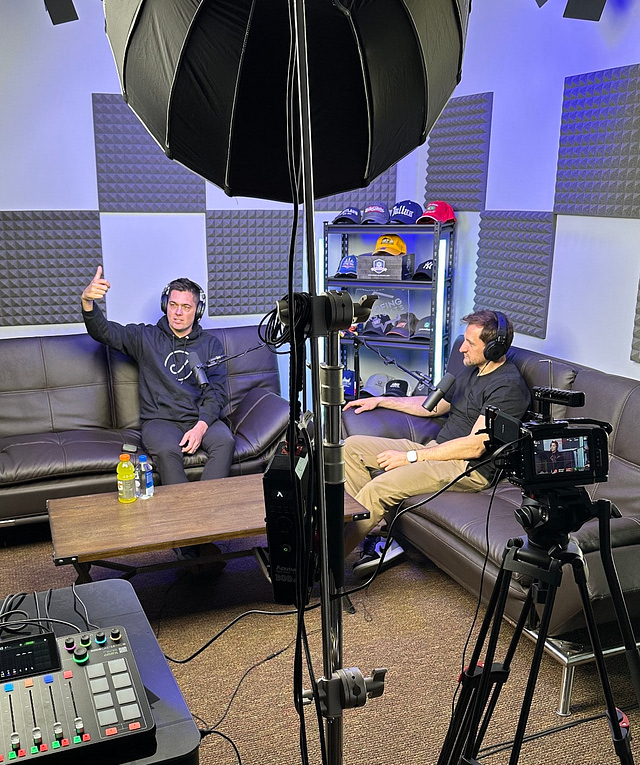 In a state-of-the-art podcast studio, two individuals wearing headphones sit on sofas. One is gesturing animatedly, sharing captivating secrets while being recorded by professional camera and audio equipment. The room is acoustically optimized with paneling and features shelves displaying a collection of caps.