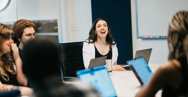 A team gathers around a conference table, focused on analyzing the new website's results. A leader in a white shirt, smiling confidently, introduces an innovative new strategy. The room is brightly lit, with a whiteboard and markers in the background.