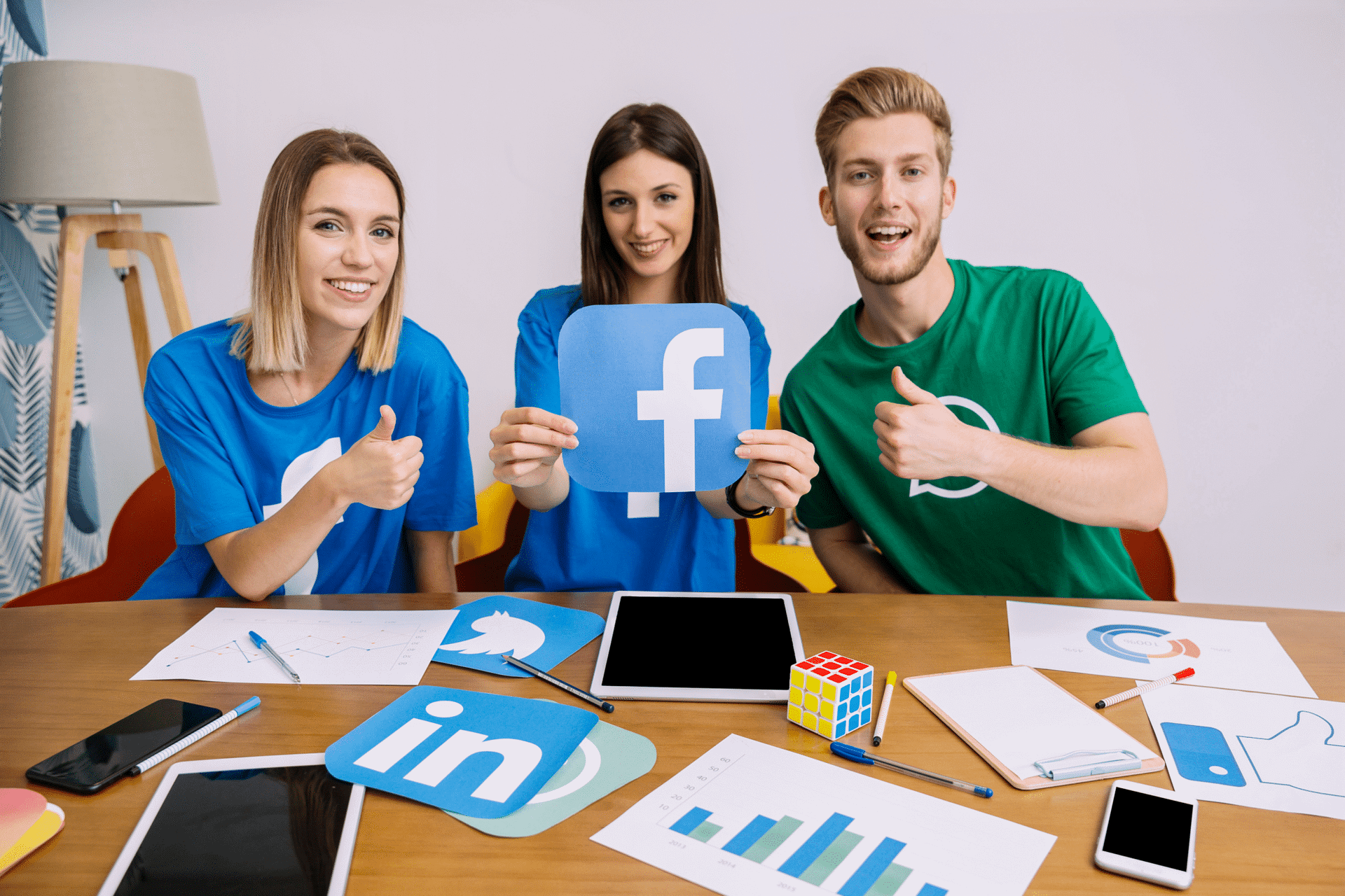 One woman holding the Facebook logo, while her 2 friends, 1 woman and 1 man are holding their thumbs up.