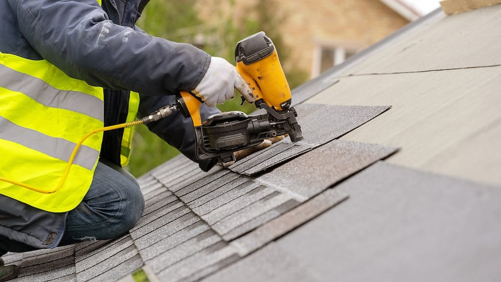 A worker wearing a neon yellow safety vest and jeans uses a nail gun to install shingles on a sloped roof